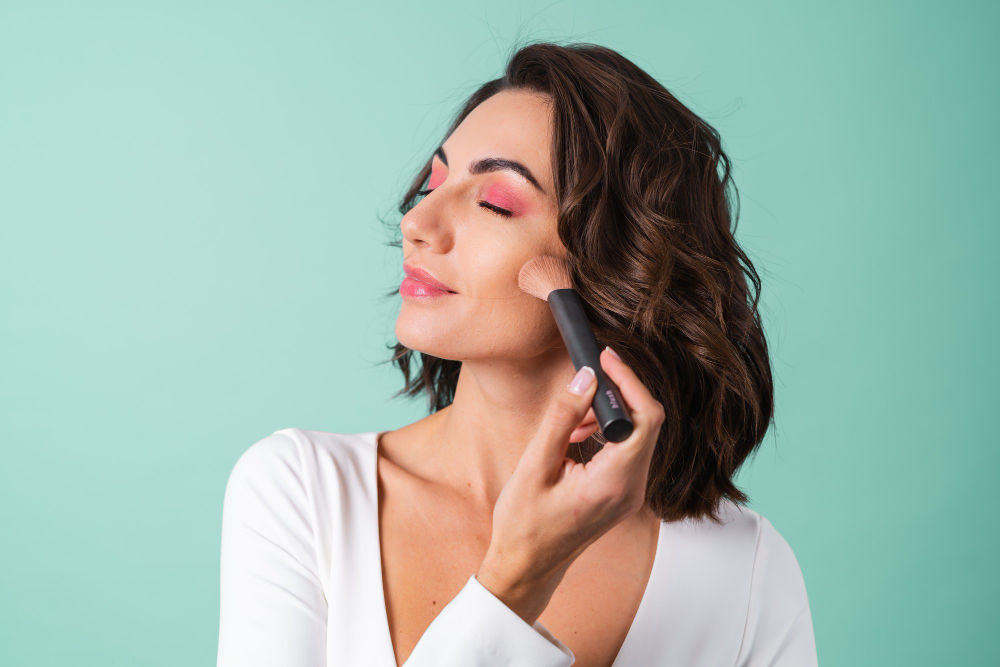 Mujer aplicándose colorete con una brocha sobre fondo verde